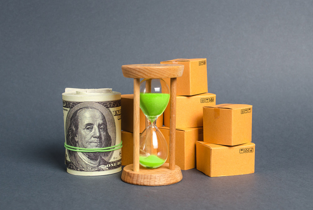 Money roll, hourglass, cardboard boxes on gray background.